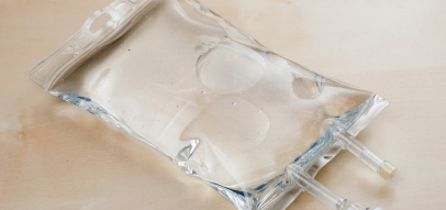 A clear intravenous (IV) fluid bag resting on a light wooden surface, featuring two attached plastic tubing for administration.