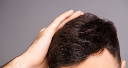 A close-up of a man touching his dark, slightly tousled hair, with a neutral gray background.