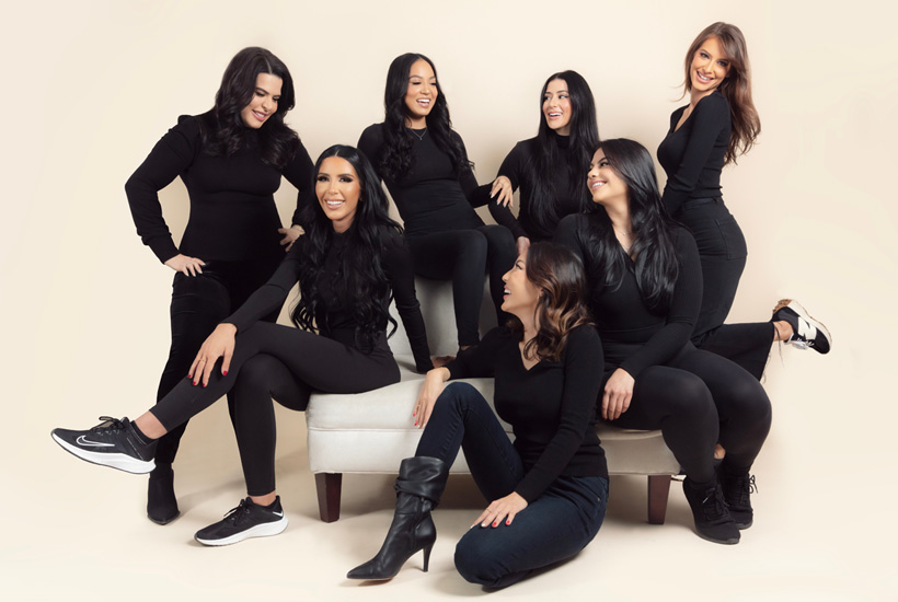 A group of women in black outfits poses on a beige background, some seated on a small sofa and others standing around.