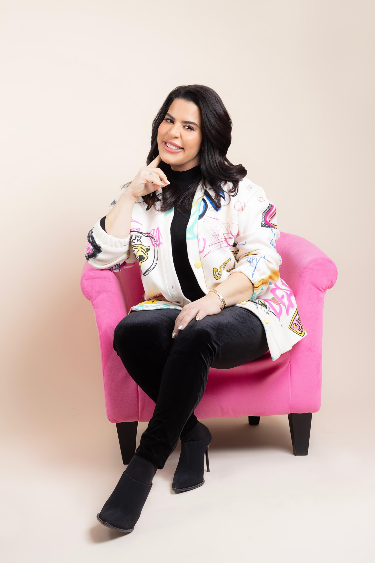 A woman wears a colorful oversized jacket and black pants, sitting confidently in a vibrant pink chair against a soft beige background.
